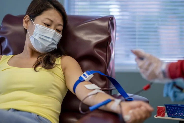 Une femme asiatique masquée d'une trentaine d'années se trouve dans un hôpital pour une collecte de sang. Elle est habillée de manière décontractée et est confortablement assise sur une chaise. Un agent de santé tient une poche de sang et prélève du sang sur son bras.